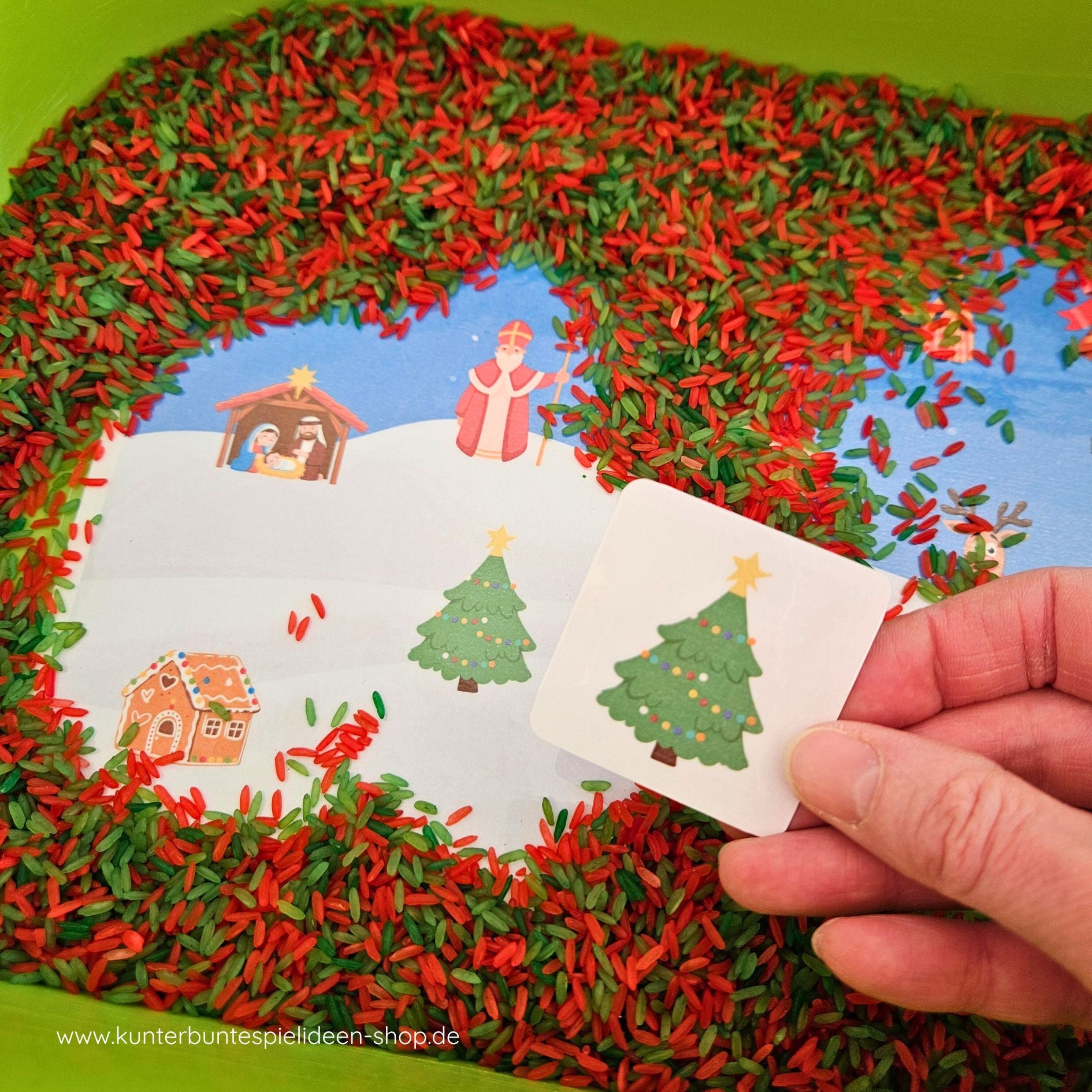 Suchen-und-Finden zu Weihnachten im Aktionstablett: Hand entdeckt den Weihnachtsbaum im rot-grünen Spielreis; weihnachtliche Symbole wie Krippe und Nikolaus sichtbar – Sensory Play als Montessori Kinderhaus-Material zur Feinmotorik und Sprachförderung (der/die/das).