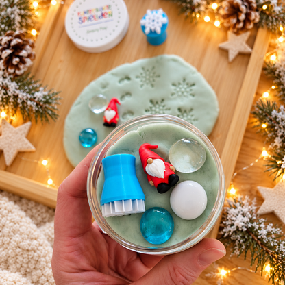 Knete in der Dose für Kinder mit rotem Wichtel, Schneeflocken-Knetstempel und drei winterlichen Muggelsteinen, in einer Hand vor weihnachtlicher Dekoration gehalten.