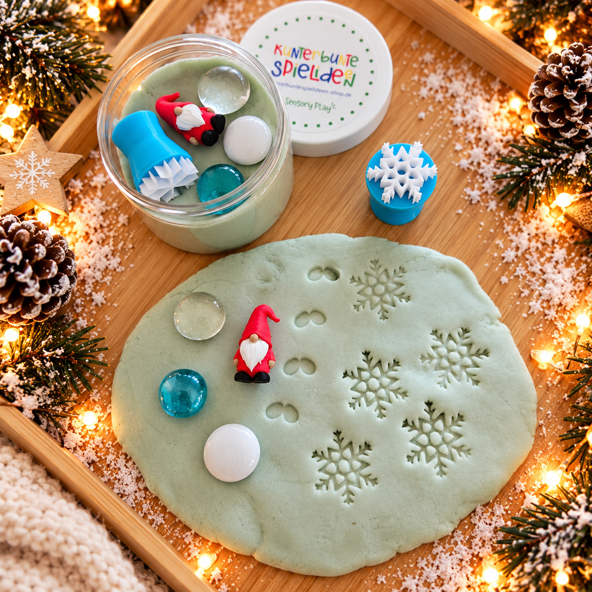 Knete in der Dose als Wichtelgeschenk für Kinder mit rotem Wichtel, Schneeflocken-Knetstempel und drei Muggelsteinen neben ausgerollter Schneeknete mit Schneeflockenabdrücken.