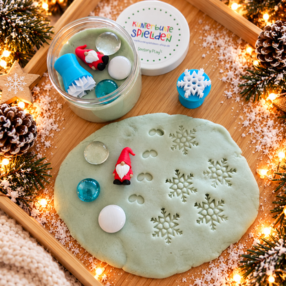Knete in der Dose als Wichtelgeschenk für Kinder mit rotem Wichtel, Schneeflocken-Knetstempel und drei Muggelsteinen neben ausgerollter Schneeknete mit Schneeflockenabdrücken.