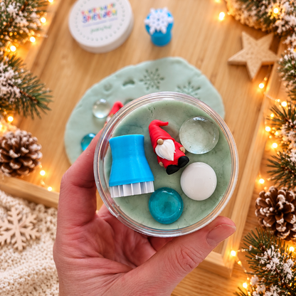 Kleine Geschenkidee für die Wichteltür: Hand hält eine Dose mit Schneeknete, roter Wichtelfigur, blauem Knetstempel und drei winterlichen Muggelsteinen.