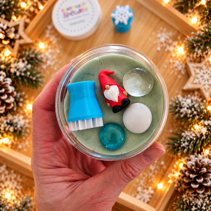 Kleine Adventskalenderfüllung mit Knete: Hand hält die geöffnete Knetdose mit Wichtelfigur, blauem Stempel und winterlichen Muggelsteinen vor einer Schneeknete-Landschaft.